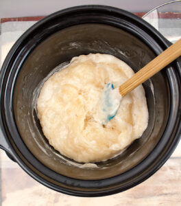 soap batter in a slow cooker with a spatula sitting in it after stirring it with a thick glossy consistency